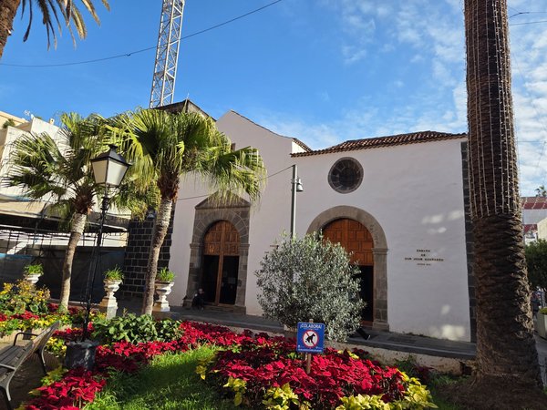 Fachada de la Ermita de San Juan Bautista en Puerto de la Cruz, con muros blancos, puertas de madera, palmeras y parterres de flores rojas en primer plano.