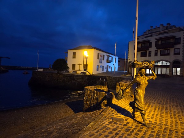 Escultura de pescadora frente al muelle y la Casa de la Real Aduana iluminada de noche en el Puerto de la Cruz.
