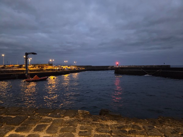 Vista nocturna del muelle del Puerto de la Cruz con luces reflejadas en el agua y el faro rojo al fondo.