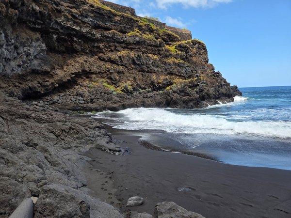 Vista de Playa Ancón: pequeña cala de arena negra volcánica con olas rompiendo junto a un acantilado de roca oscura en un día despejado.