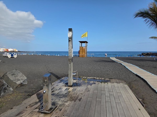 Ducha de playa y pasarela de madera en Playa Jardín de Puerto de la Cruz, con arena volcánica negra y bandera amarilla ondeando junto al mar.